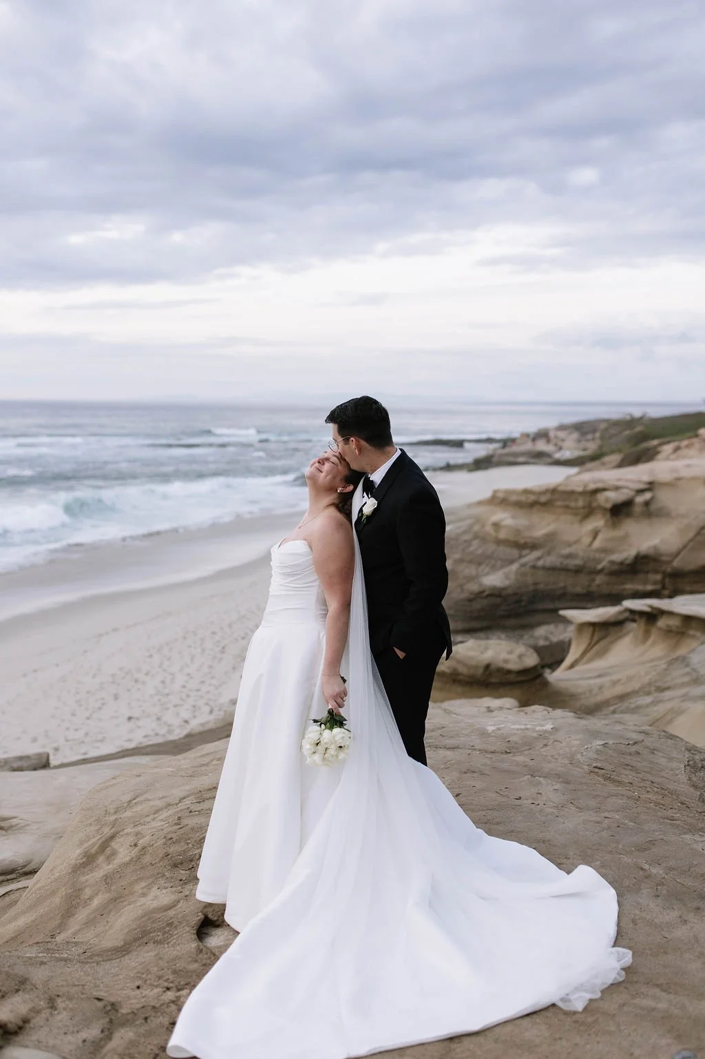 A bride and groom in wedding attire on a rocky beach, sharing a kiss with the ocean and cloudy sky in the background.