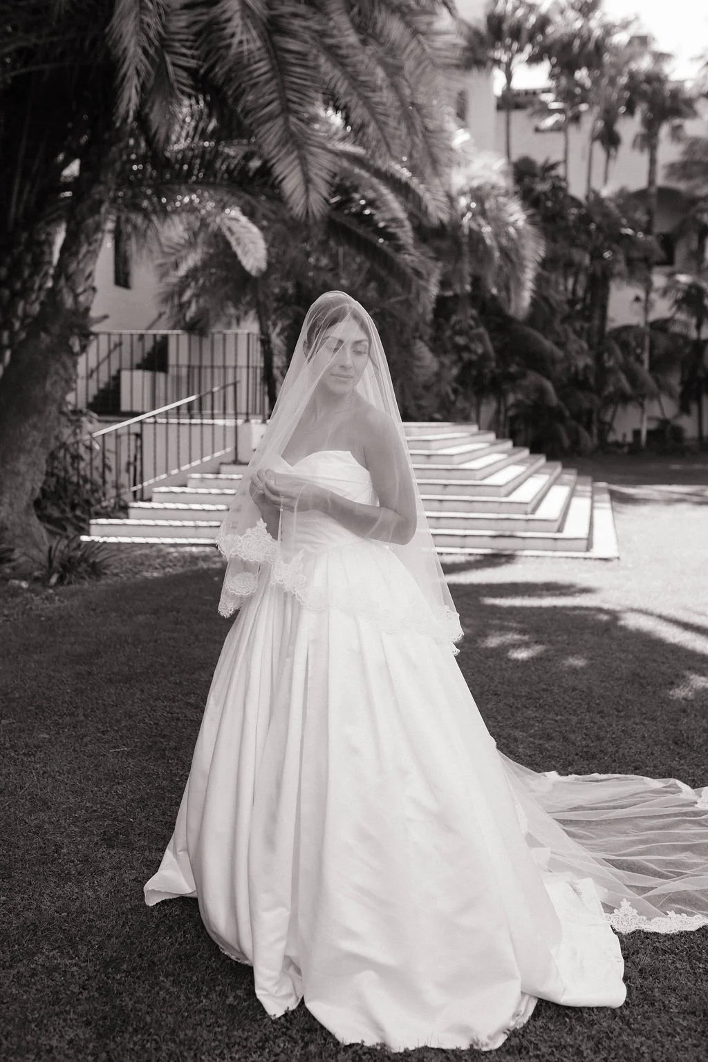A bride in a white wedding gown and veil standing outdoors on grass, with tropical plants and stairs in the background.