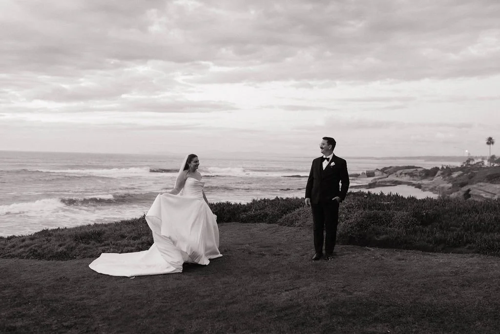 A bride and groom in wedding attire standing on a grassy area near the ocean at sunset, with cliffs and palm trees in the background.