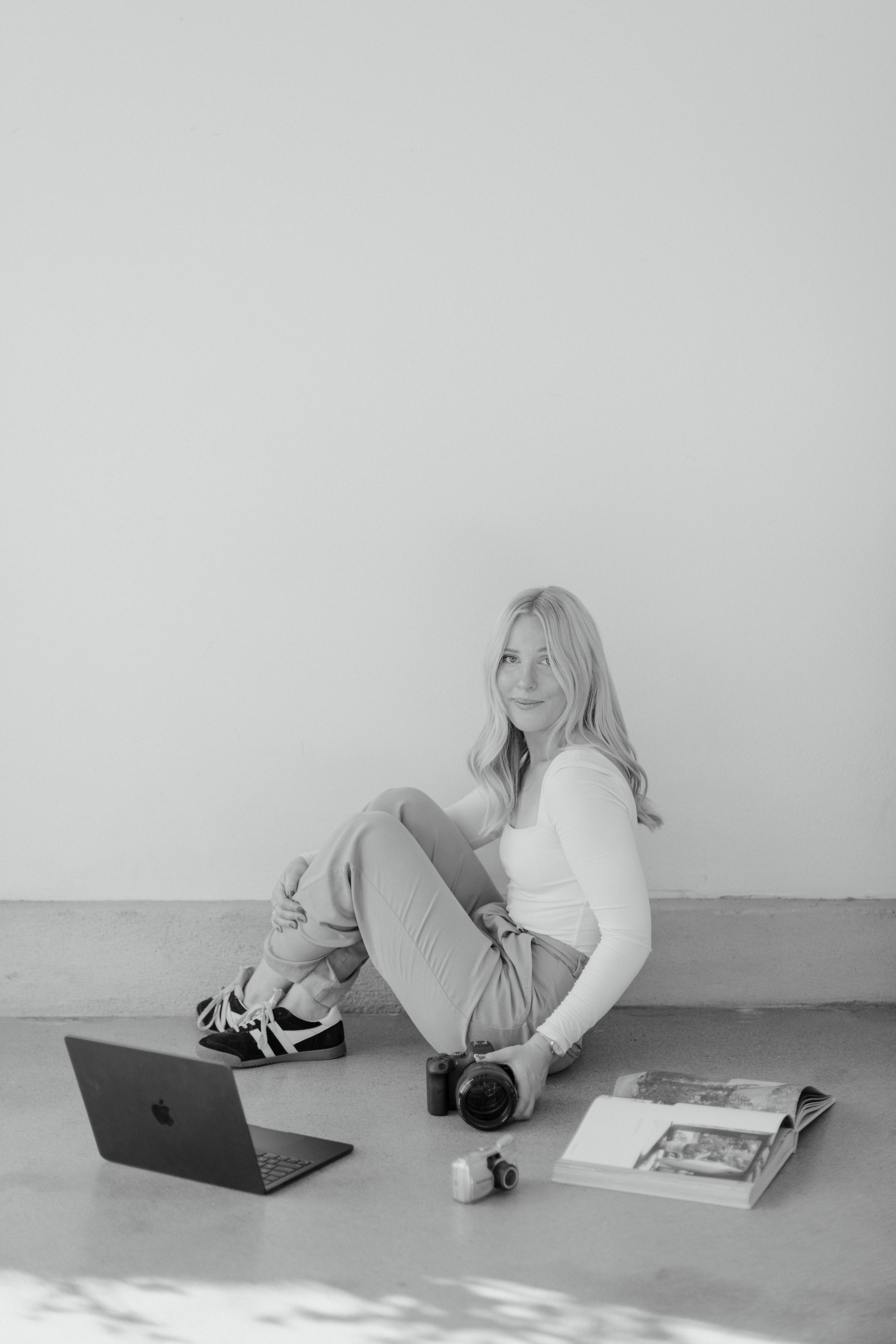 A woman with blonde hair sitting on the floor, surrounded by a laptop, camera, small camera, and open photo album, in a minimalist setting with a plain wall background.