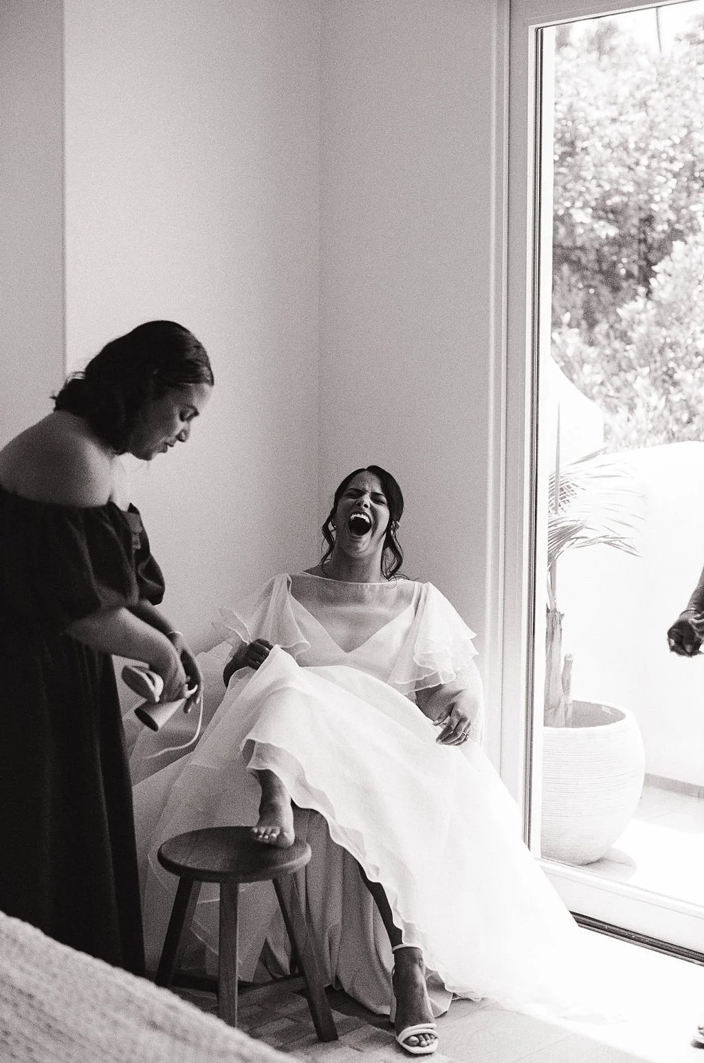 A bride is sitting on a chair in a wedding dress, laughing with her mouth open, while someone nearby styles her hair. There's a large window next to her showing outdoor plants.