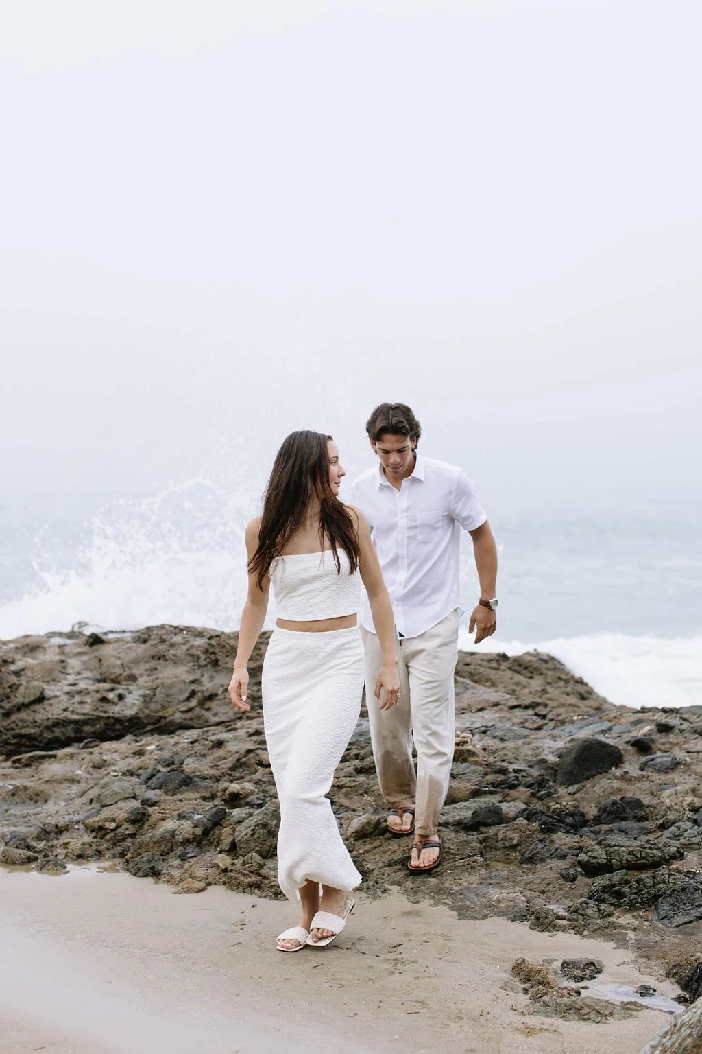 A young woman and man walk on a rocky beach near the ocean, dressed in white casual clothing, with gray overcast sky and waves in the background.