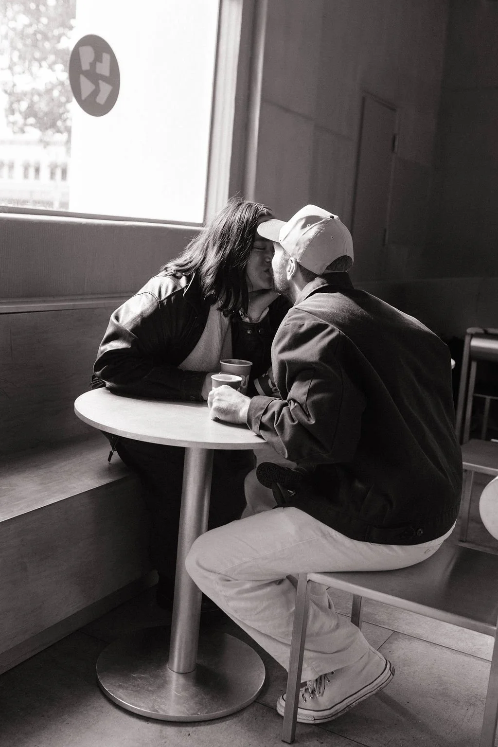 A black and white photo of a couple kissing at a small round table in a cafe. The woman has shoulder-length hair and is wearing a jacket, while the man is wearing a cap, a dark jacket, and light-colored pants.