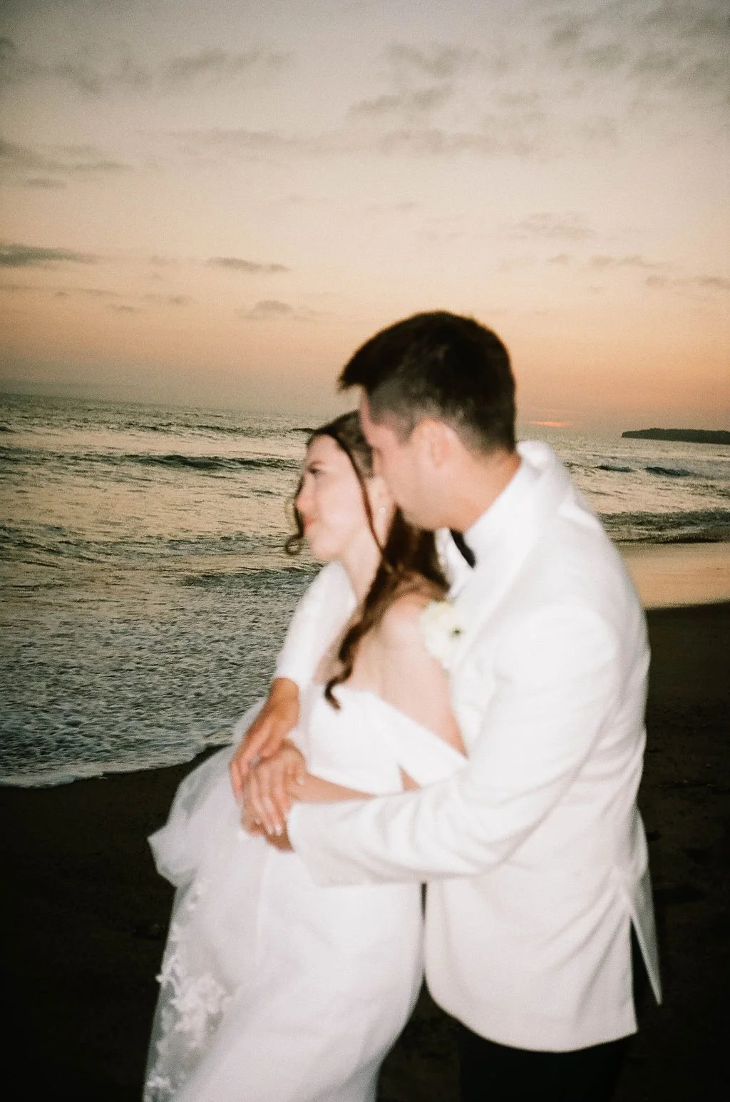 A bride and groom in white wedding attire embrace on a beach at sunset, with ocean and sky in the background.