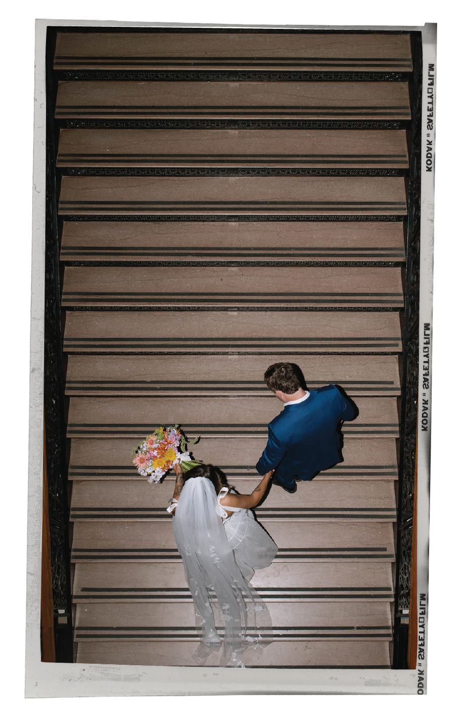 A bride and groom walking down a staircase, with the bride holding a bouquet of flowers and the groom dressed in a blue suit, viewed from above.
