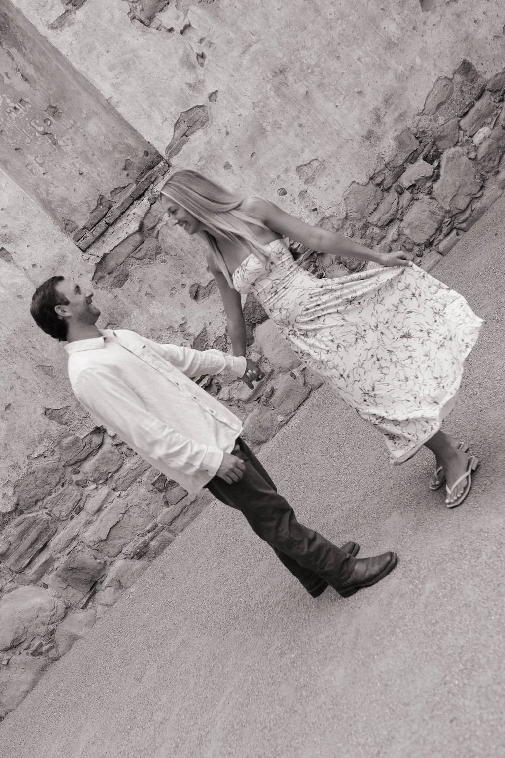 A couple holding hands and smiling at each other, standing outdoors near a weathered stone wall.