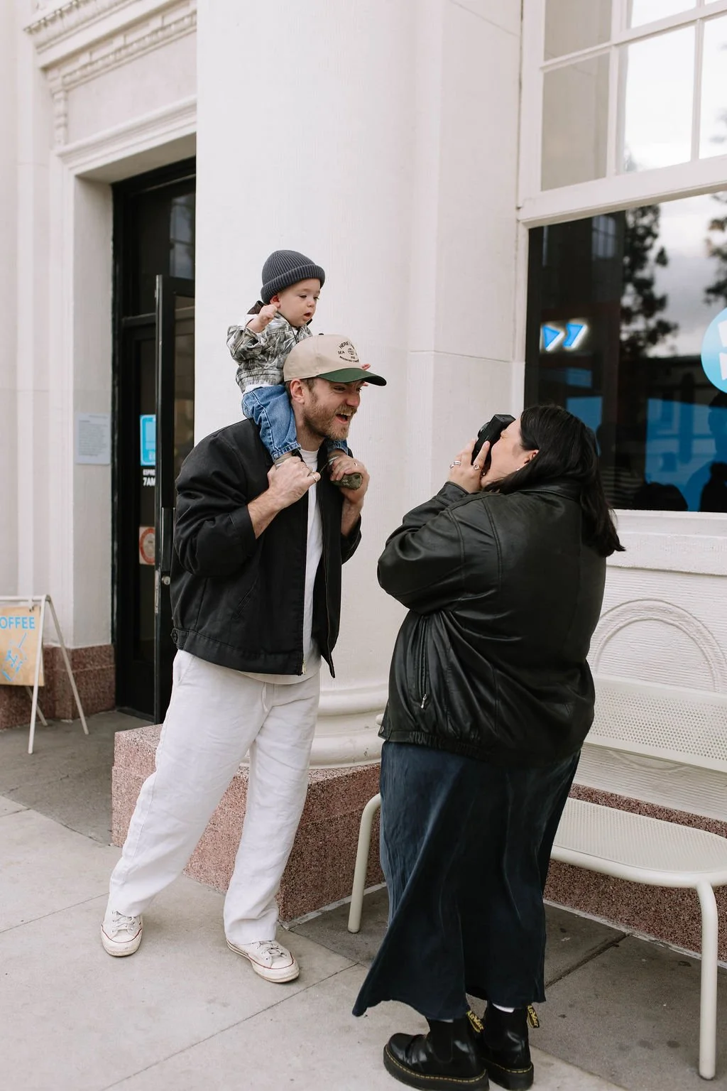 A man with a child on his shoulders talking to a woman who is taking their picture outside a building.