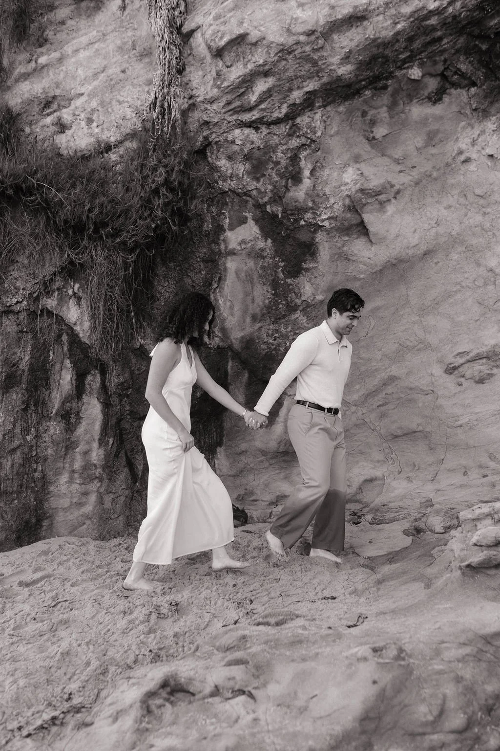 A couple holding hands walking on sandy terrain near a rocky cliff. The woman is wearing a long, sleeveless dress, and the man is dressed in a long-sleeve shirt and trousers, both barefoot. The scene is in black and white.