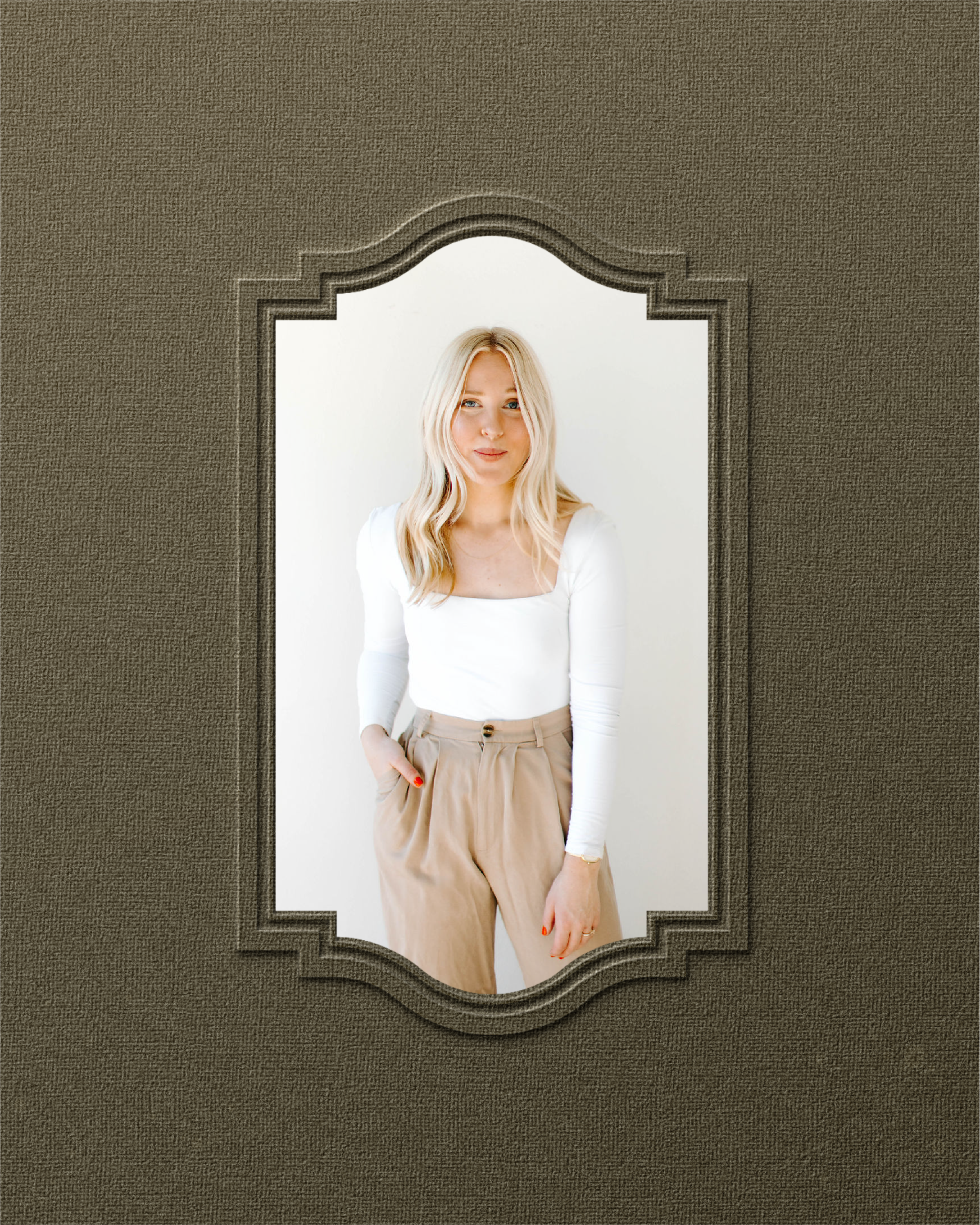 A woman with blonde hair wearing a white long-sleeve top and beige high-waisted pants, standing against a white background, viewed through a textured brown picture frame.