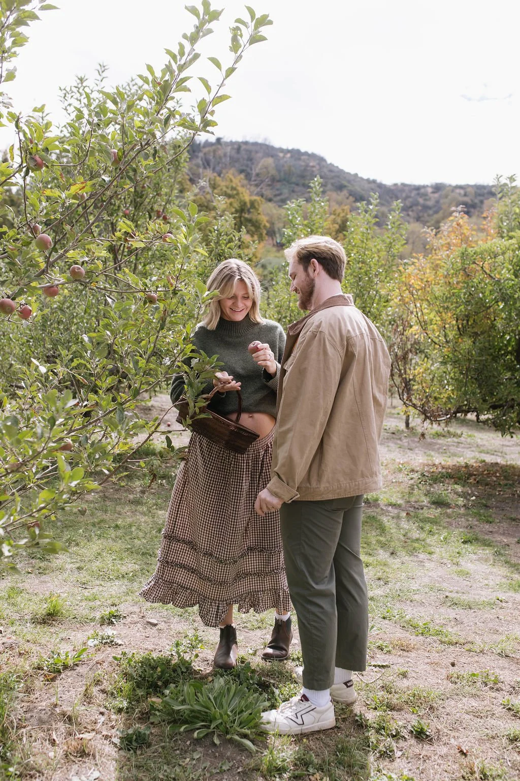 A young woman and man, enjoying a day at an apple orchard, talking and smiling near apple trees with mountains in the background.
