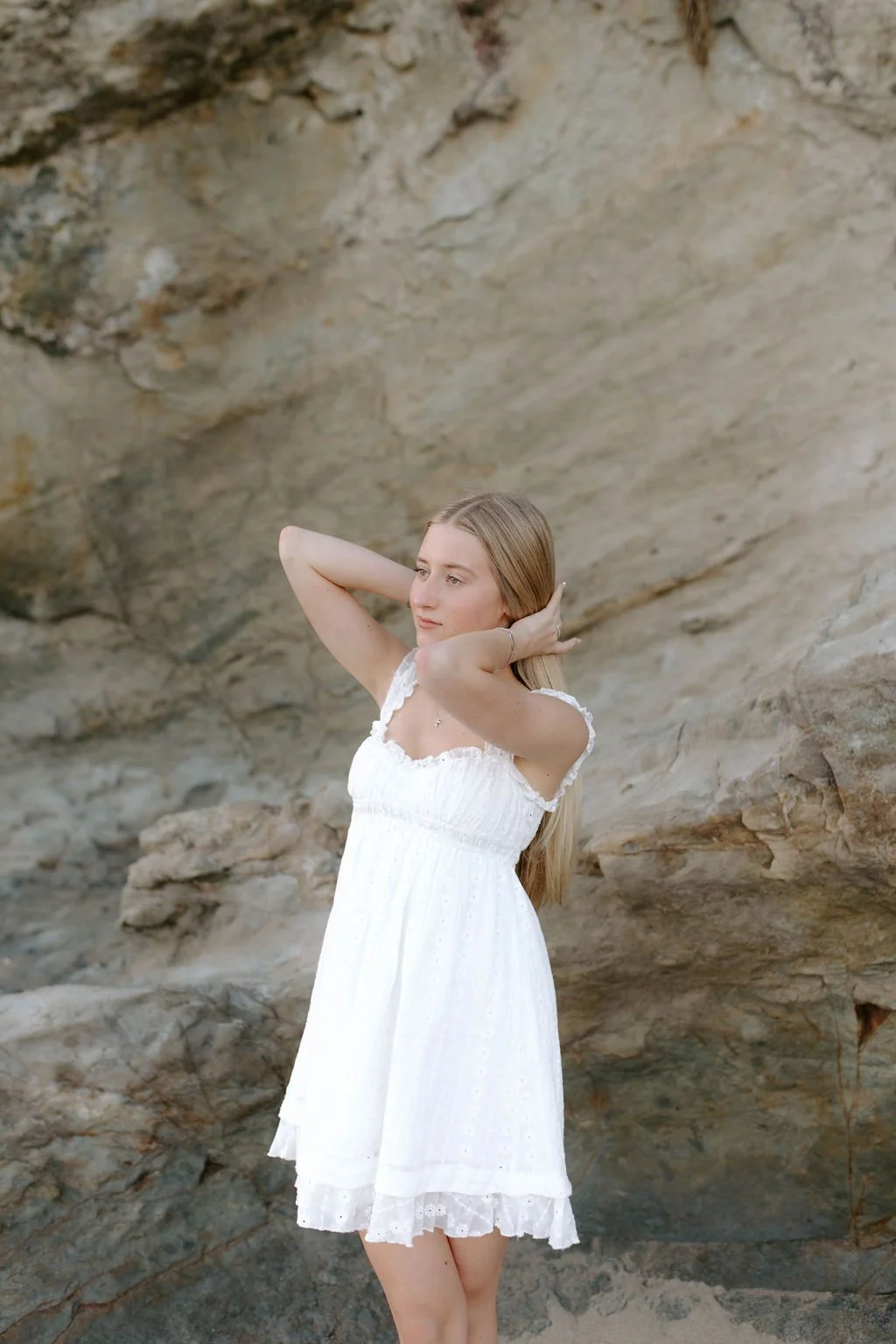 A young woman with long blonde hair wearing a white dress, standing near a rocky cliff with her hands behind her head, looking off into the distance.