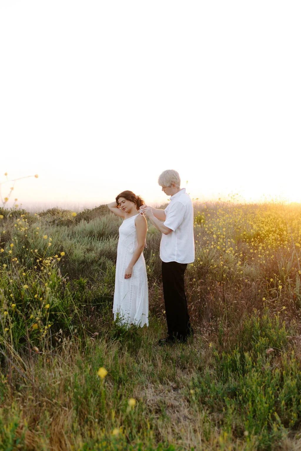 A man with silver hair in a white shirt is fixing a woman's dress in a field of yellow wildflowers during sunset.