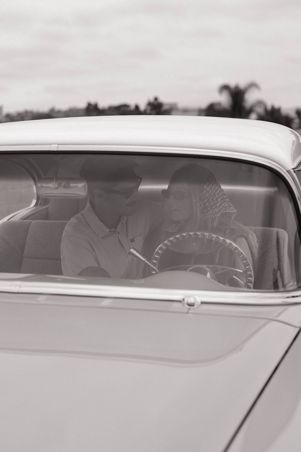 A young man and woman sitting close together inside a vintage car, leaning in as if sharing a moment, with the man in a hat and the woman wearing glasses and a headscarf.