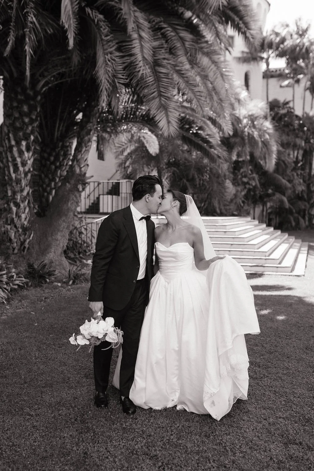 A bride and groom kiss outdoors, the groom holding a bouquet, surrounded by palm trees and stairs in the background.