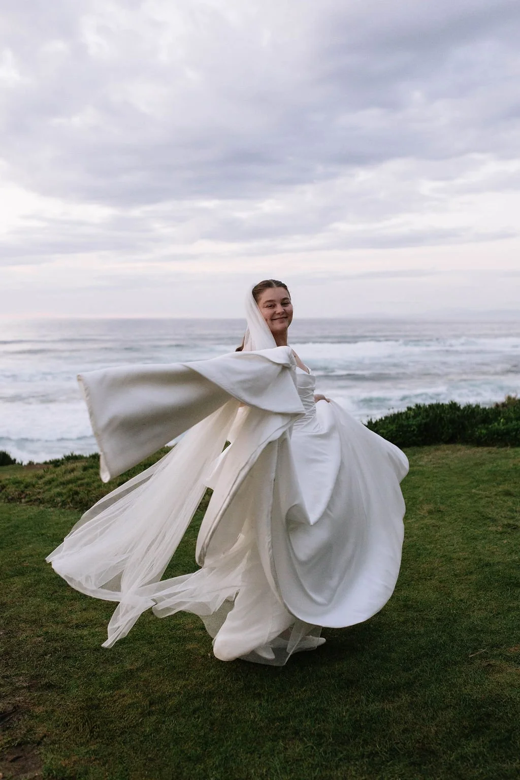 A smiling bride in a flowing white wedding dress dancing on a grassy area near the ocean with a cloudy sky overhead.
