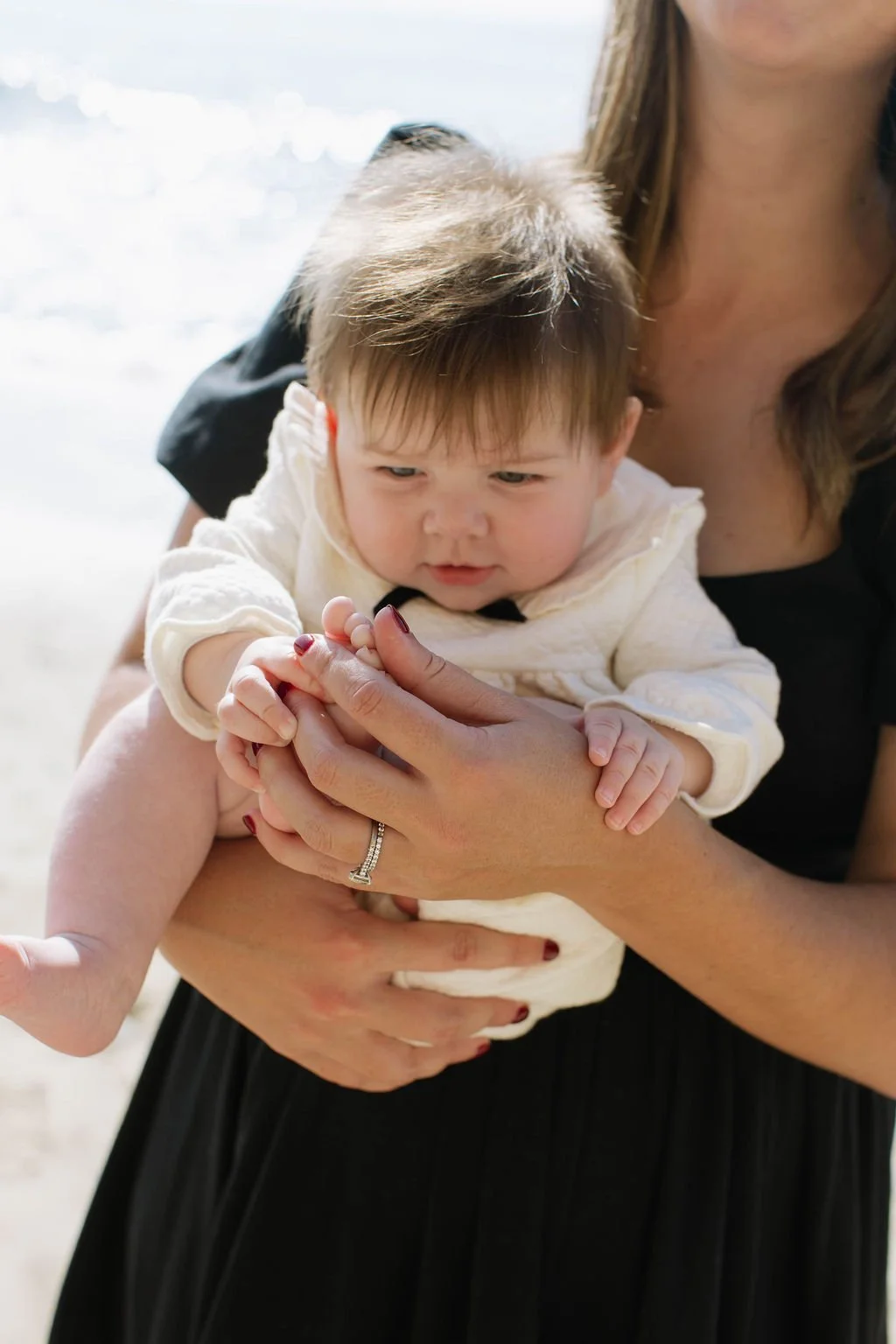 A woman holding a young child in her arms at the beach, with the ocean in the background. The child is looking at her hand, which the woman is holding.