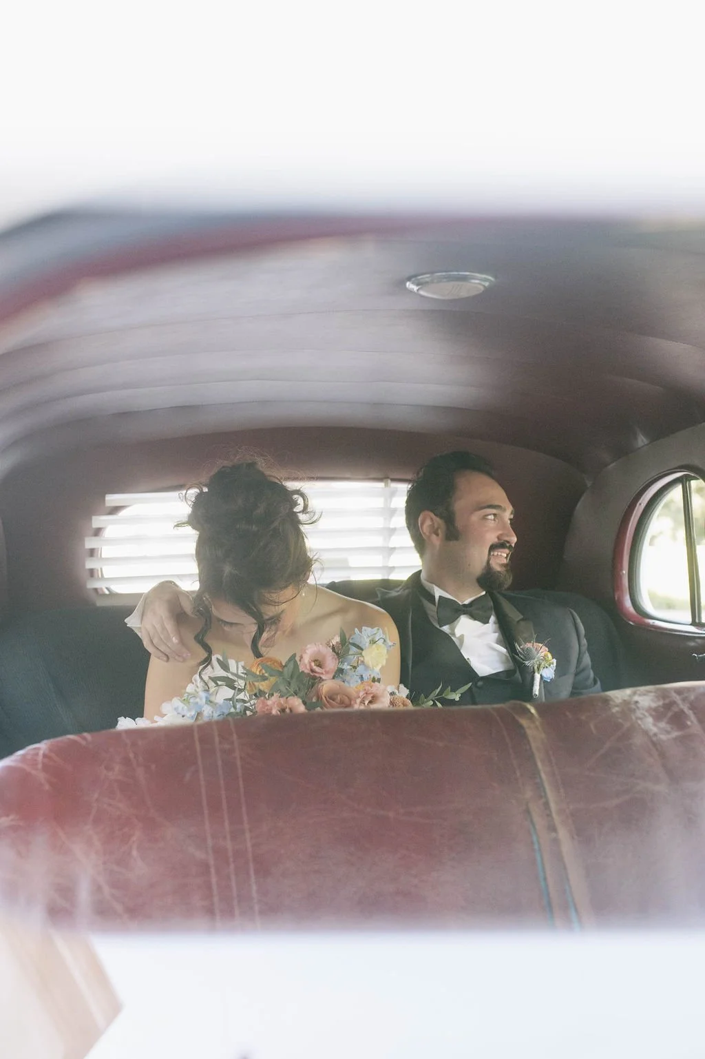 A bride and groom sitting in the back of a vintage car, with the bride leaning on the groom, holding a bouquet of flowers, both dressed in wedding attire, inside the car.