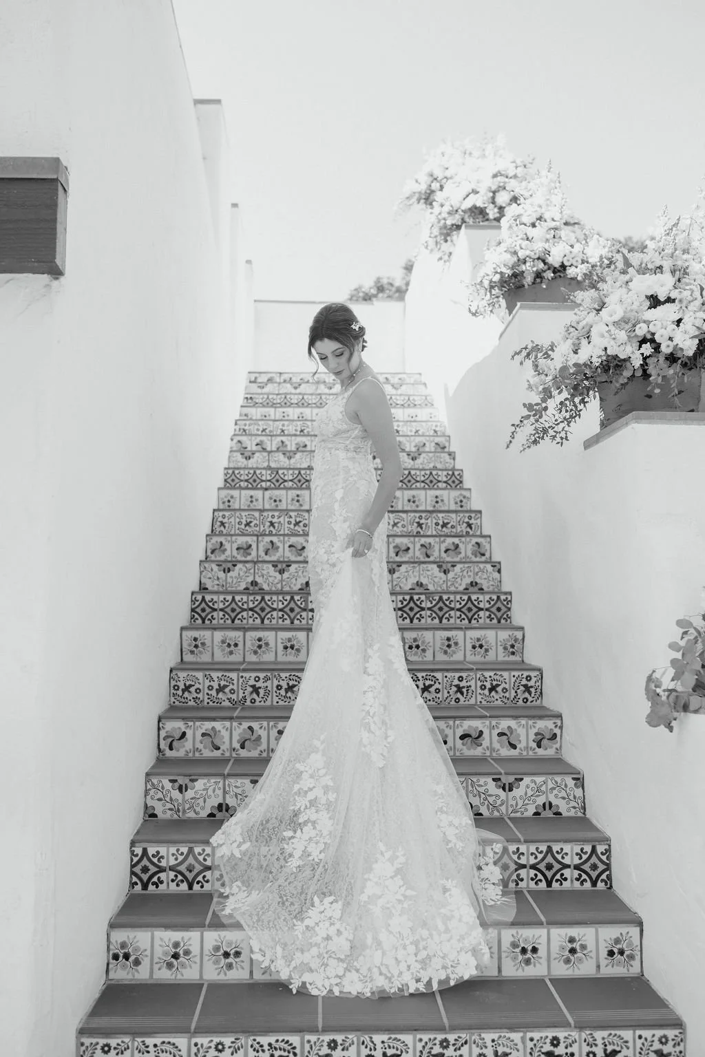 Black and white photo of a bride in a lace wedding gown standing on decorative tiled stairs, surrounded by white walls and large flower pots with blooming flowers.