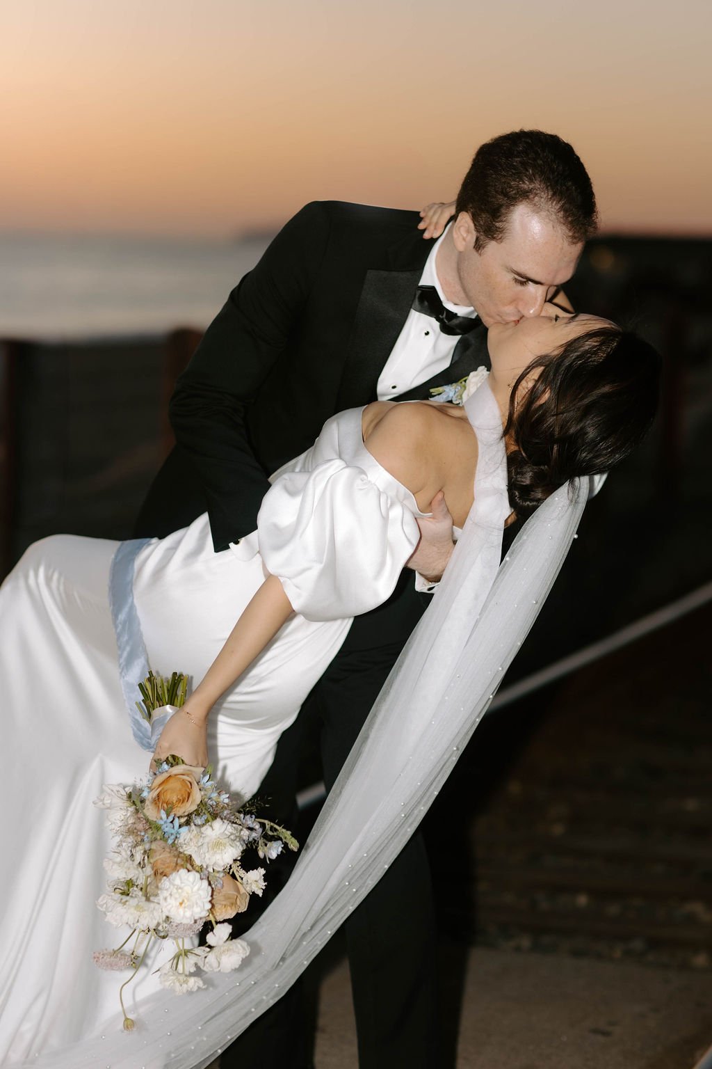 A groom in a black tuxedo and a bride in a white wedding dress sharing a kiss on the beach during sunset.