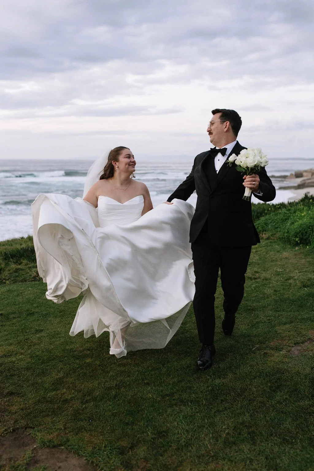 A bride and groom on a grassy area near the ocean on their wedding day. The bride is wearing a strapless white wedding gown and the groom is in a black tuxedo, holding a bouquet of white flowers.