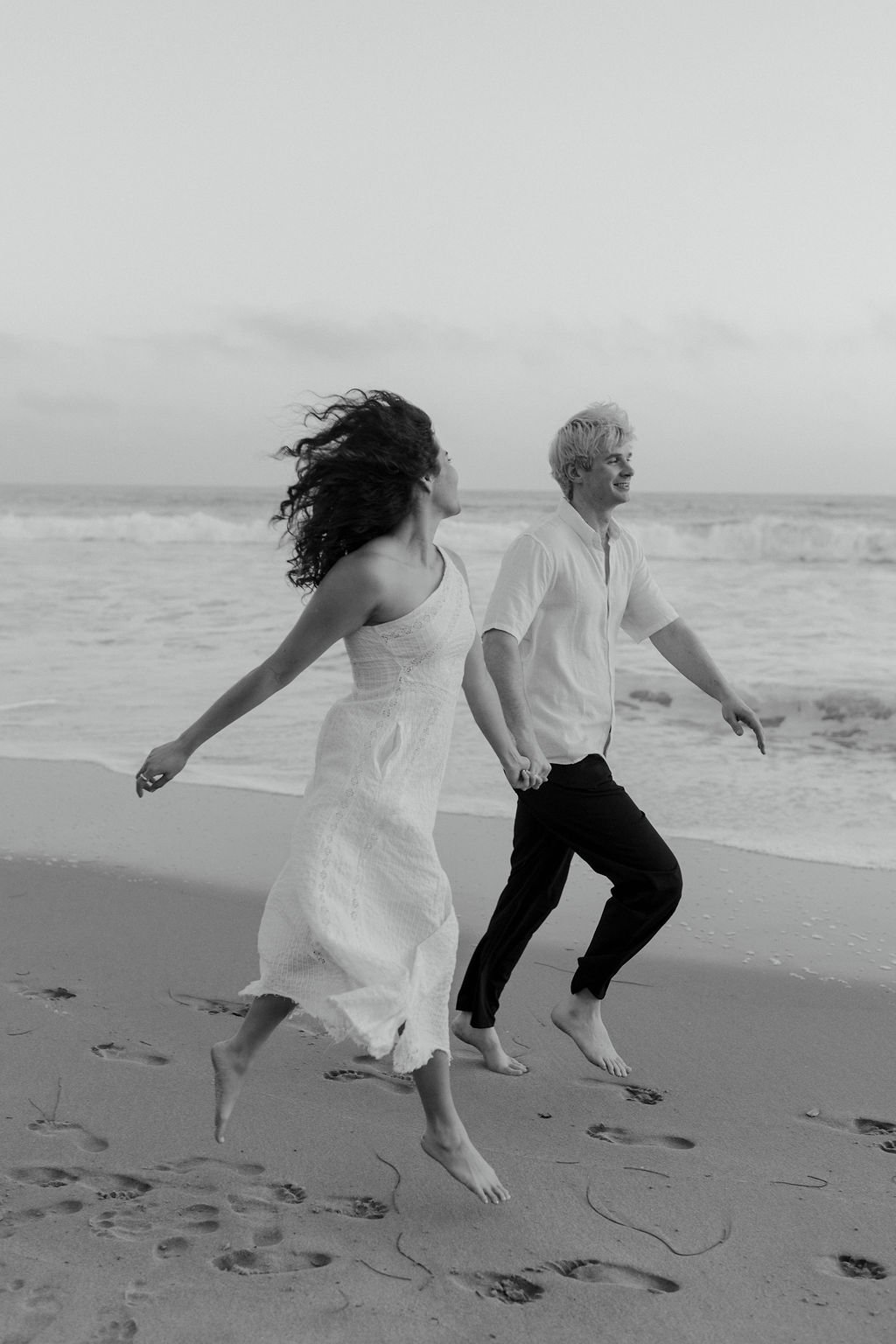 A black-and-white photo of a couple running barefoot on the beach, holding hands, with the ocean waves in the background.