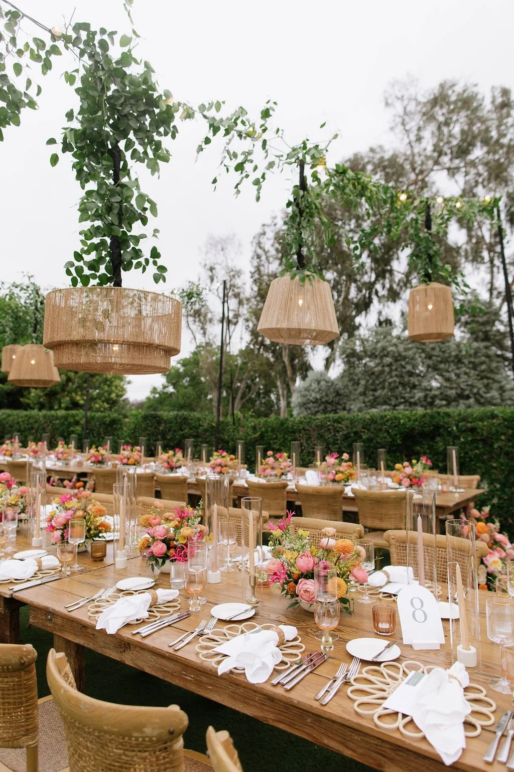 A beautifully decorated outdoor dining setup with long wooden tables, floral centerpieces, and hanging woven lamps, set amidst greenery.