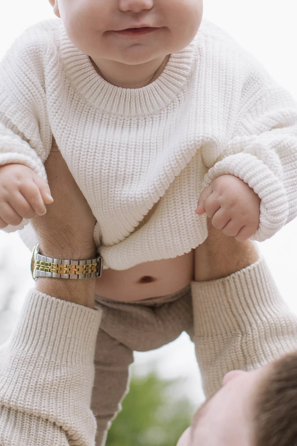A young child being held up by an adult outdoors, wearing a cream-colored sweater and beige pants, with a focus on the child's upper body and face.