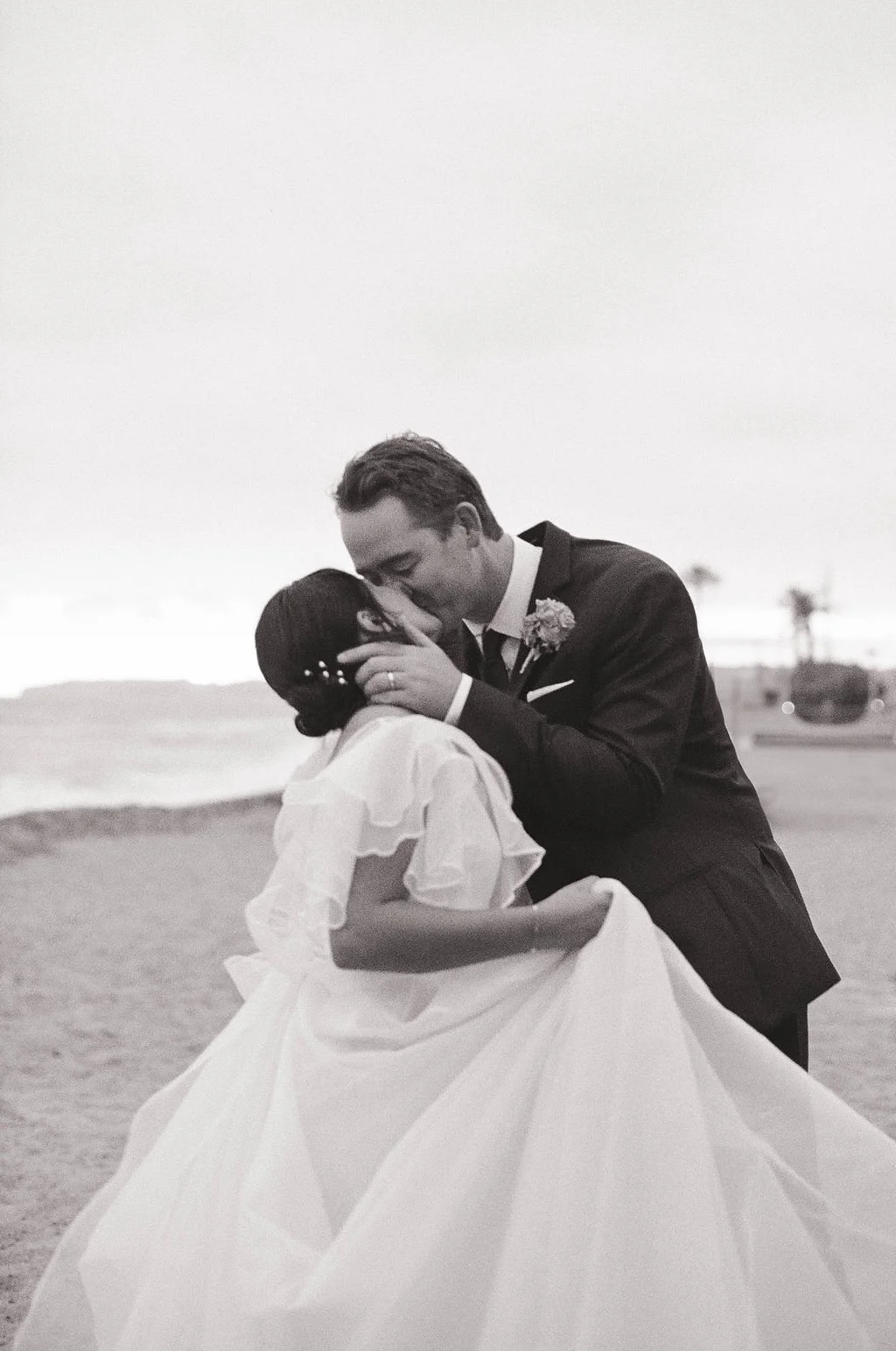 A black and white photo of a newlywed couple sharing a kiss on the beach, with the groom in a suit and the bride in a white wedding dress.