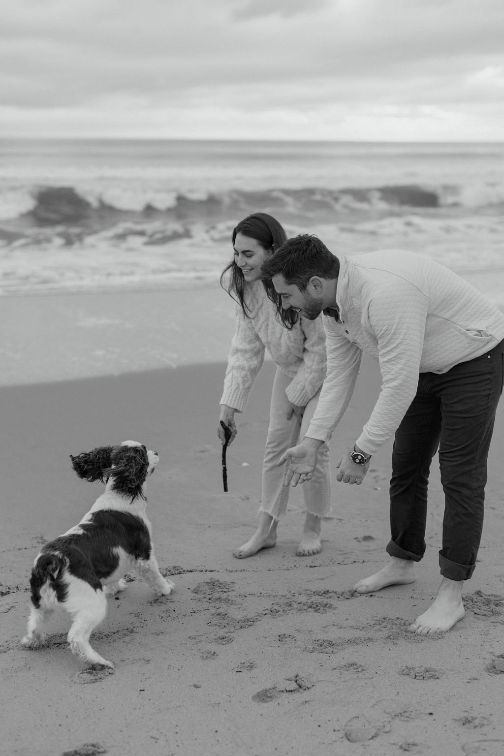 A couple and their dog playing with a stick on the beach, with ocean waves in the background, black and white image.