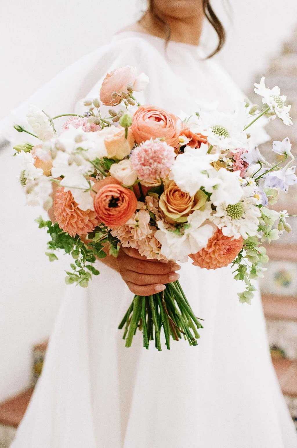 A person holding a large bouquet of pink, white, and peach flowers, with a person wearing a white dress visible in the background.