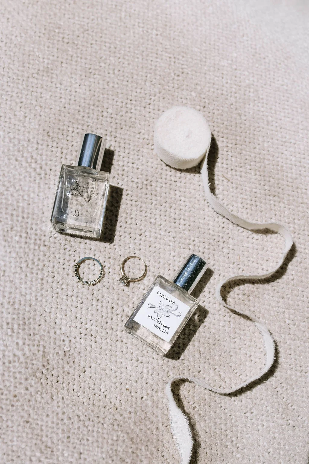 Two bottles of perfume, two rings, a white makeup sponge, and a white ribbon are arranged on a textured beige surface.