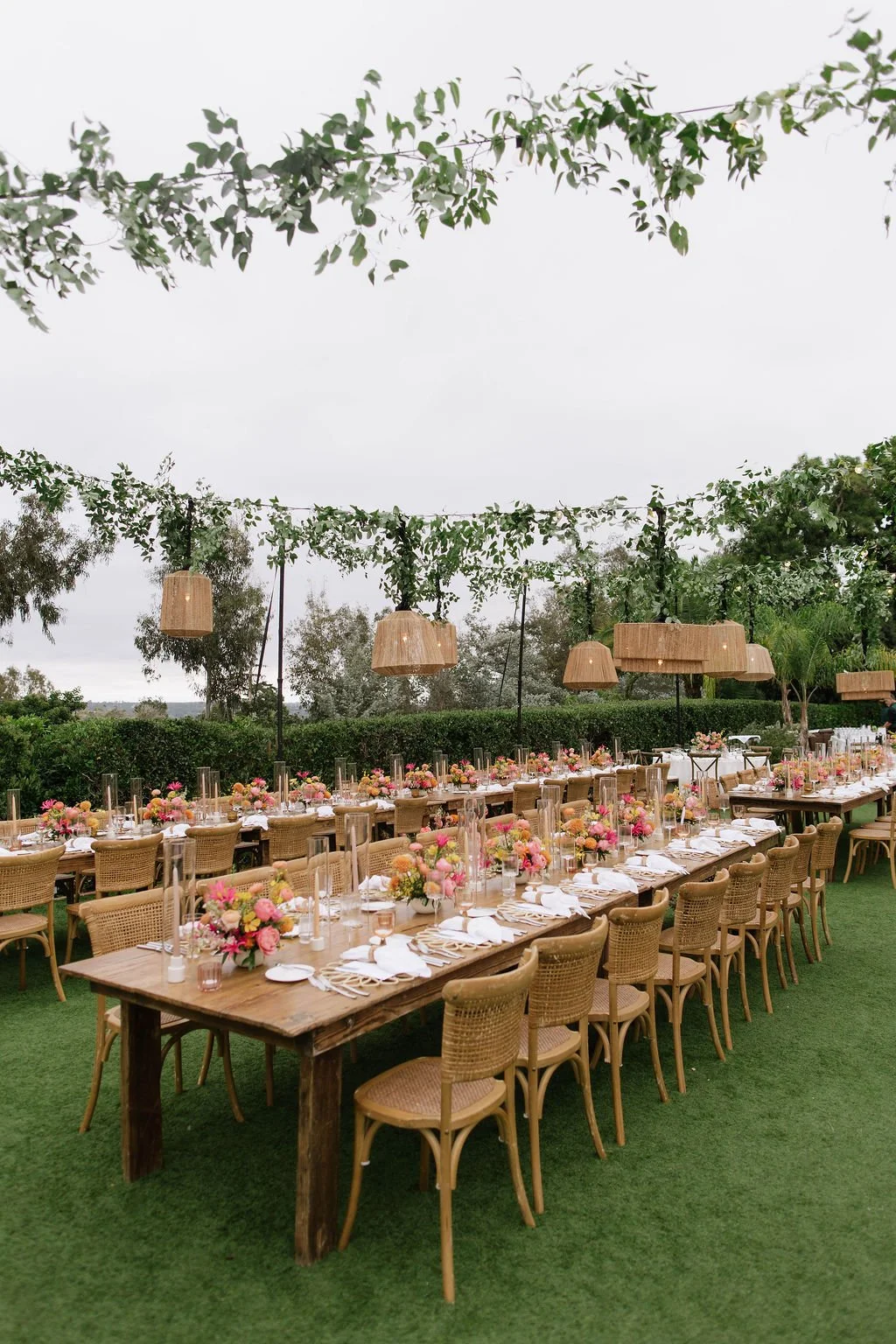 Outdoor wedding reception setup on a lawn with long wooden tables, pink and peach floral centerpieces, white plates, and glassware, under hanging wicker lamps and green leafy garlands, with trees and hedge in the background.