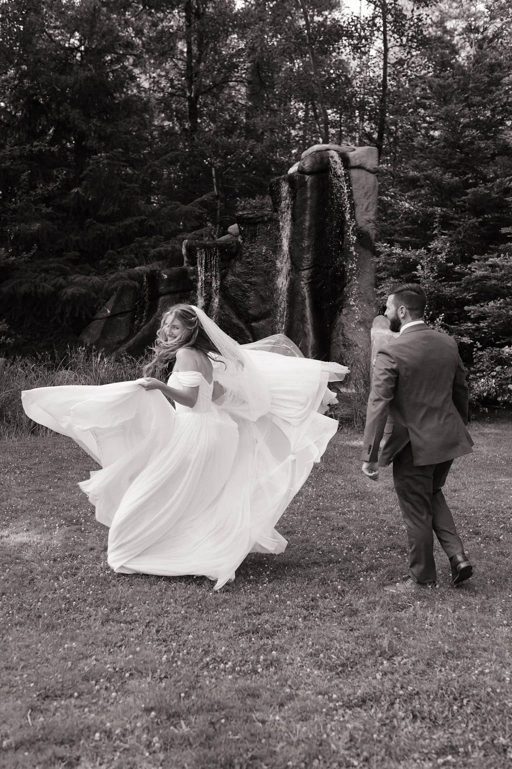 A bride in a flowing white wedding dress and veil twirls playfully, smiling, as a groom in a suit follows her on a grassy area with a waterfall and trees in the background.