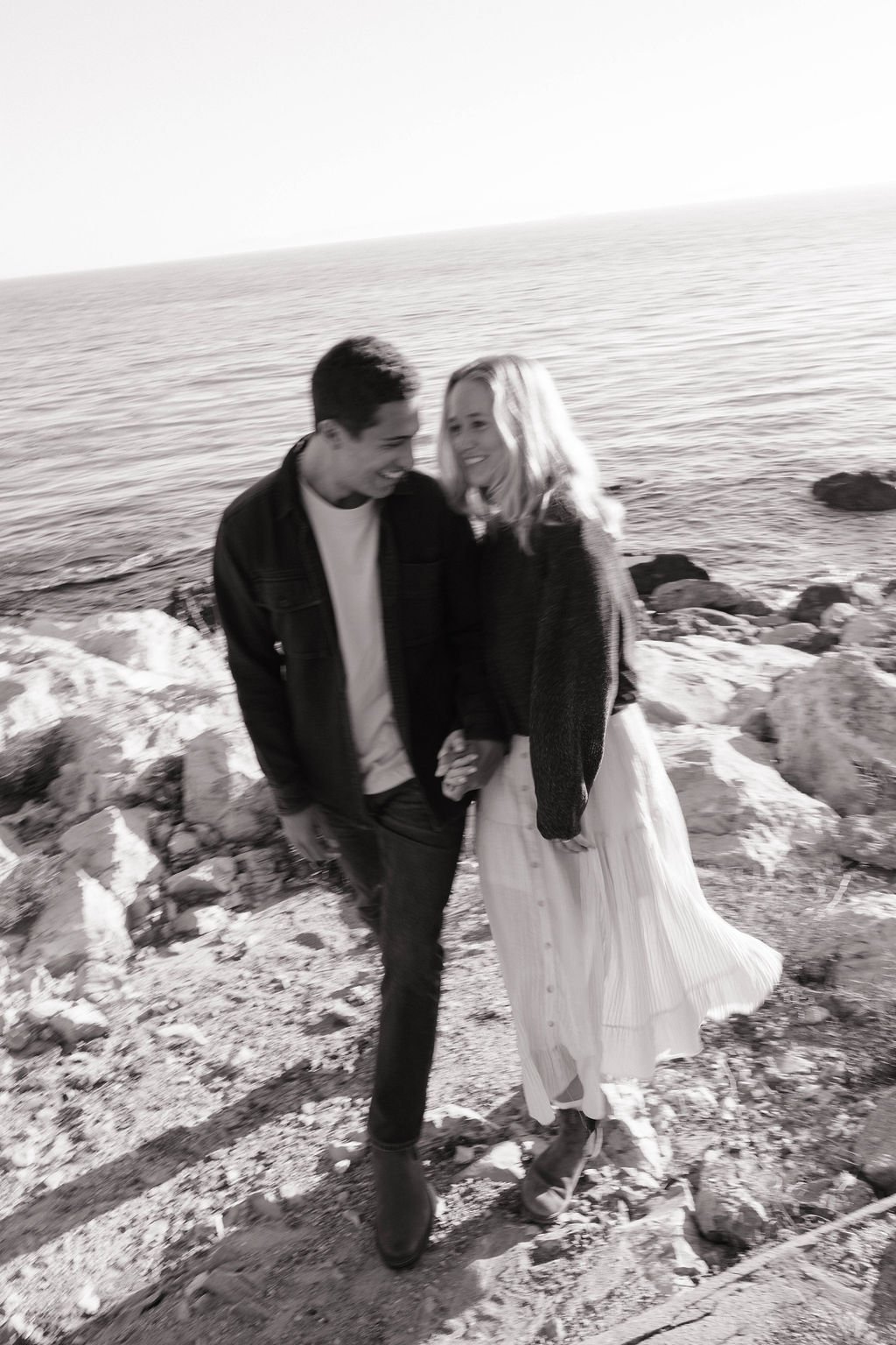 A black and white photo of a young couple holding hands and smiling at each other on a rocky beach near the ocean.