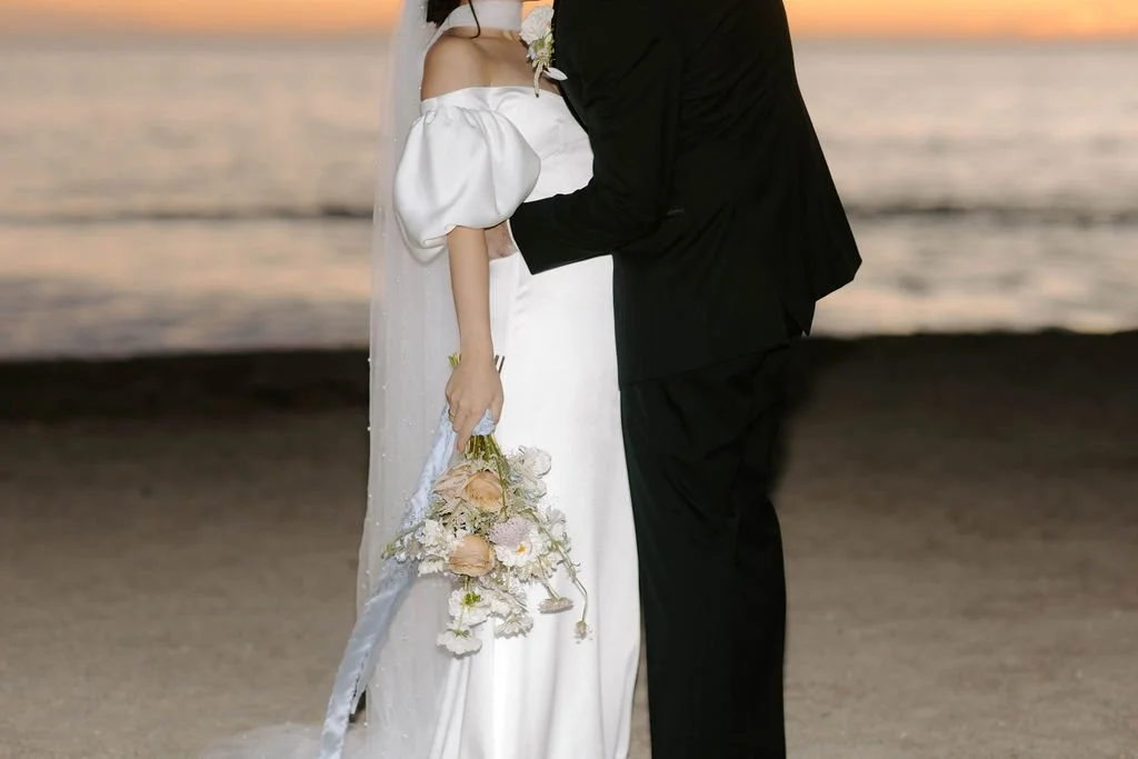 A bride and groom stand close together on a beach at sunset, with the ocean in the background. The bride is wearing an off-the-shoulder white wedding dress with puffy sleeves, and is holding a bouquet of pale flowers. The groom is dressed in a black 