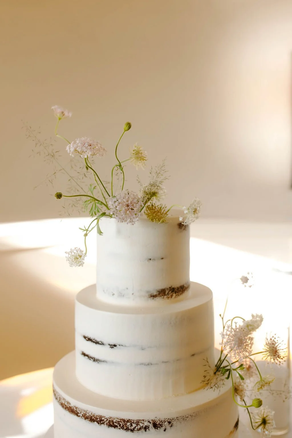 A three-tier wedding cake with white frosting and minimal design, decorated with light pink and white flowers and green stems.
