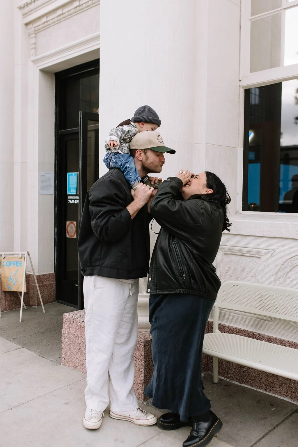 A family of three, a man, woman, and a young boy, sharing a joyful moment outside a building, with the boy sitting on the man's shoulders.