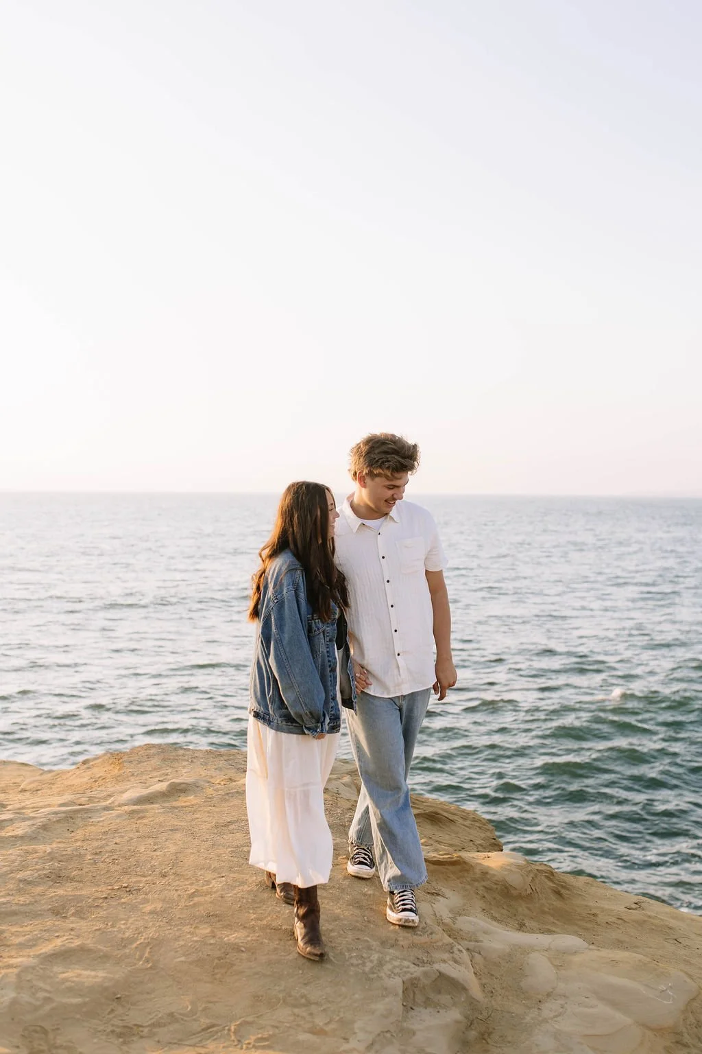 A young couple walking hand-in-hand on a rocky shoreline by the ocean with clear skies.