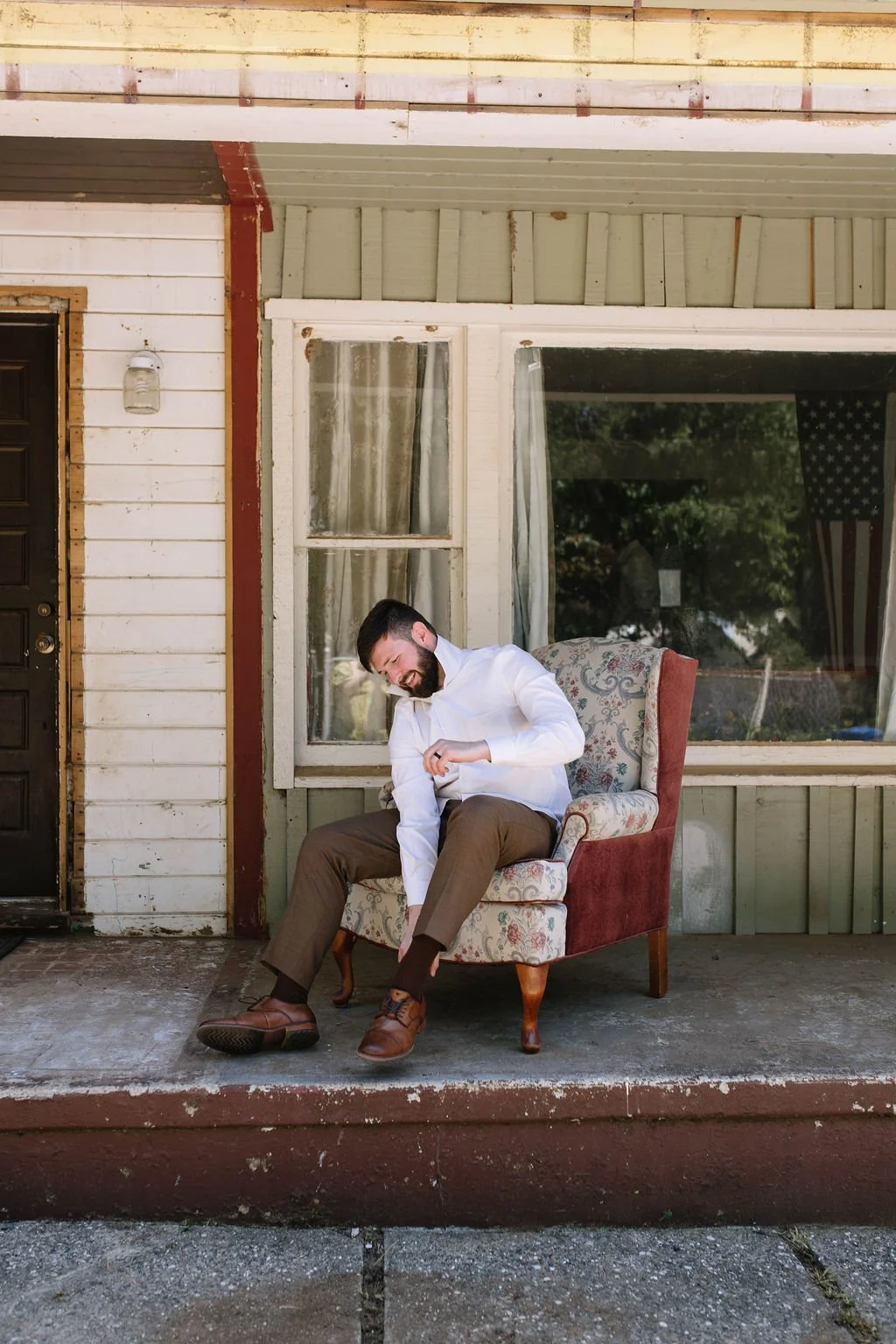 A man sitting on a vintage floral armchair outside a house, laughing and adjusting his sock.