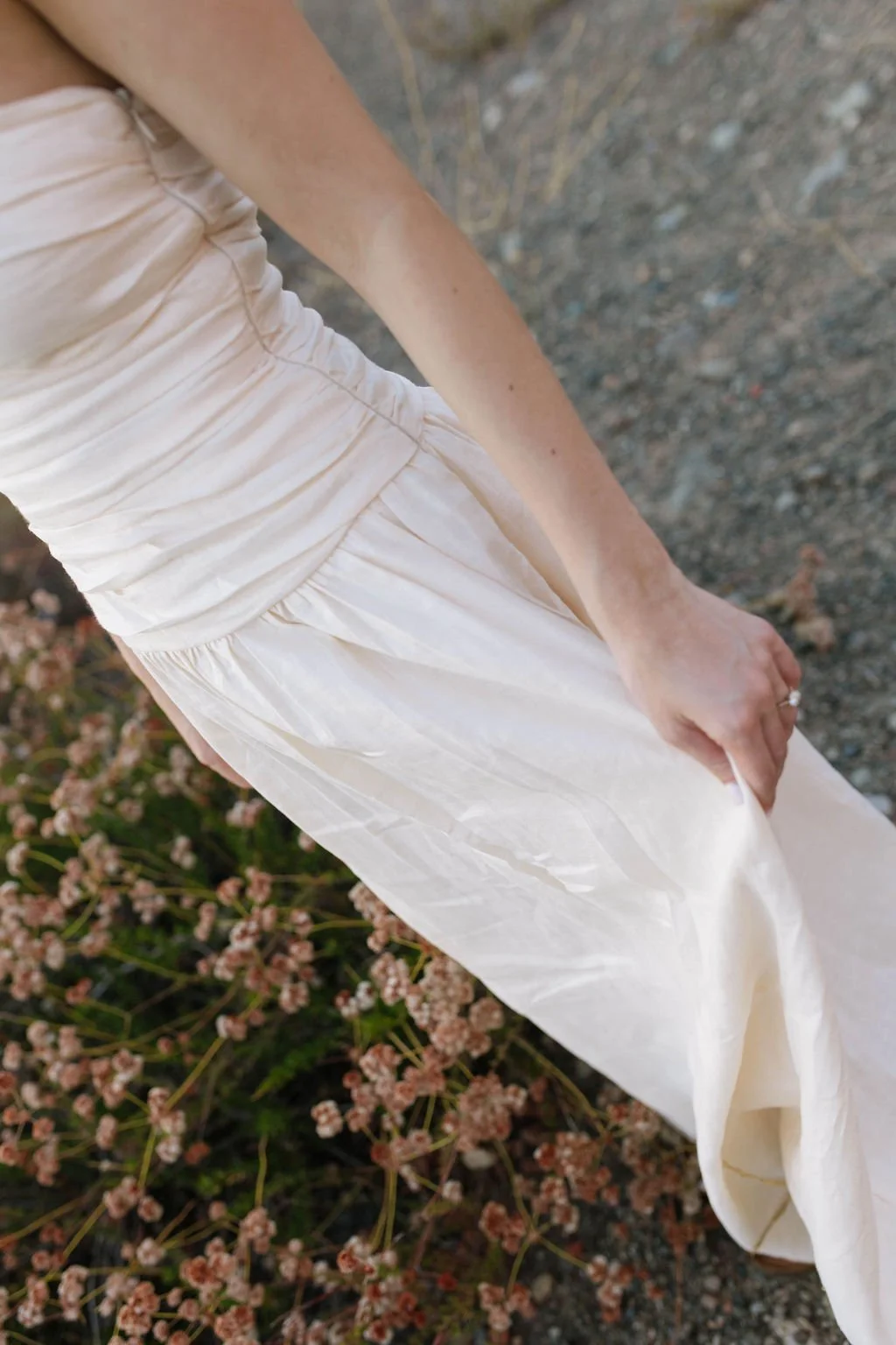 A person wearing a white dress or gown, holding the fabric as they walk or stand outdoors on a gravel path, with small pink flowers nearby.