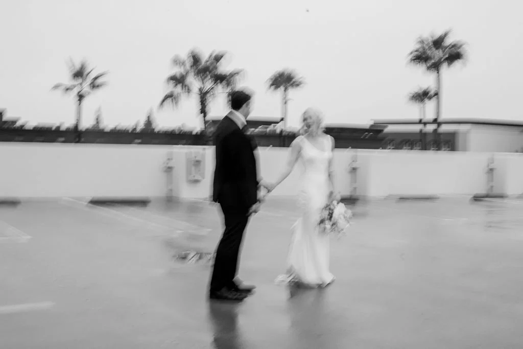 A black and white photo of a bride holding a bouquet and a groom standing on a rooftop parking lot, with palm trees and a cloudy sky in the background.