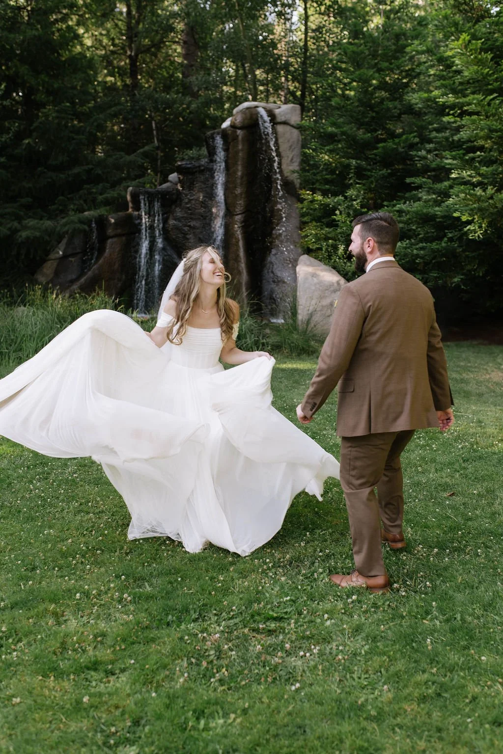A bride and groom in wedding attire dancing outdoors on grass, with a waterfall and lush greenery in the background.