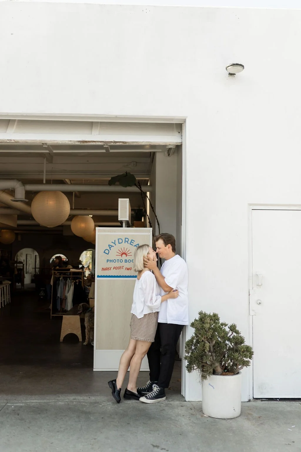A couple standing outside a photo booth, about to kiss, in front of a white wall and a sign that says 'Daydream Photo Booth'.