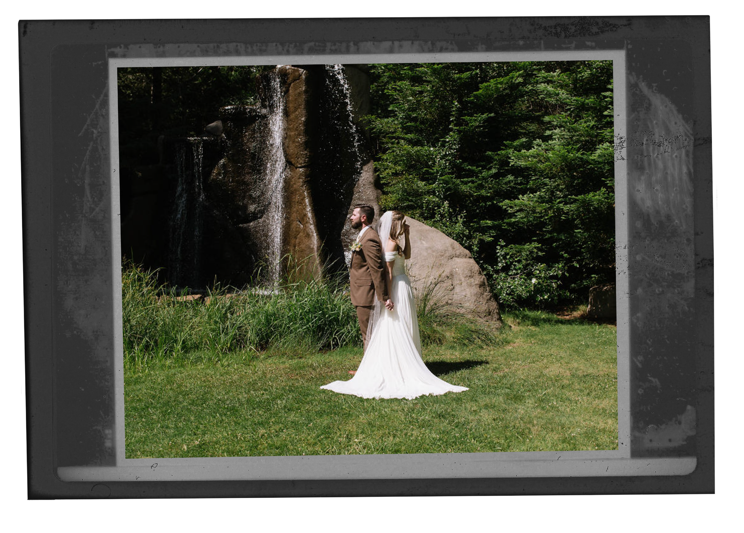 A bride and groom stand back to back outdoors on grass, holding hands behind their backs near a large rock and waterfall, surrounded by greenery.