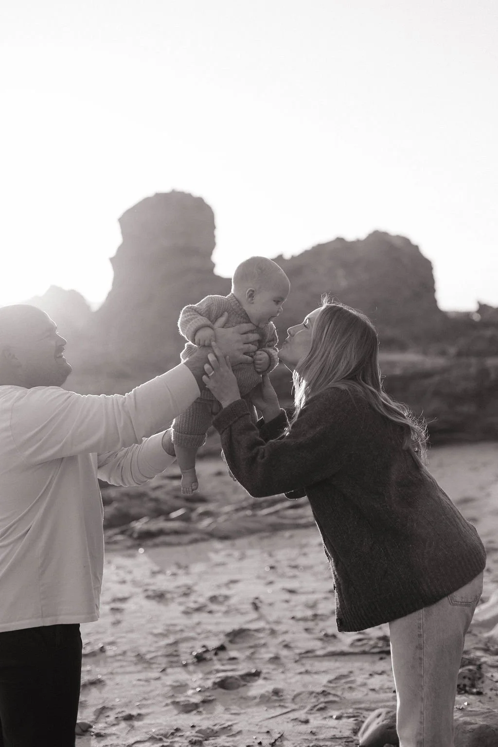 A joyful family on the beach with rocky formations in the background, where a man and woman are playing with their baby.