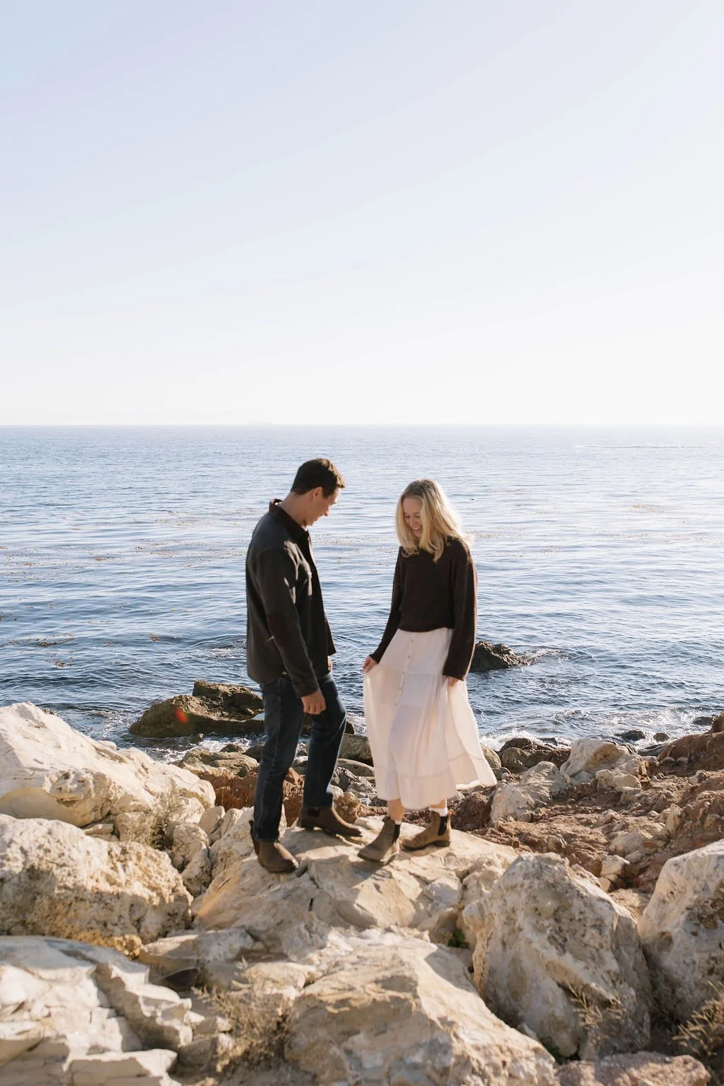 A young man and woman standing on rocks by the ocean, smiling and looking down at their feet, with the sea and sky in the background.