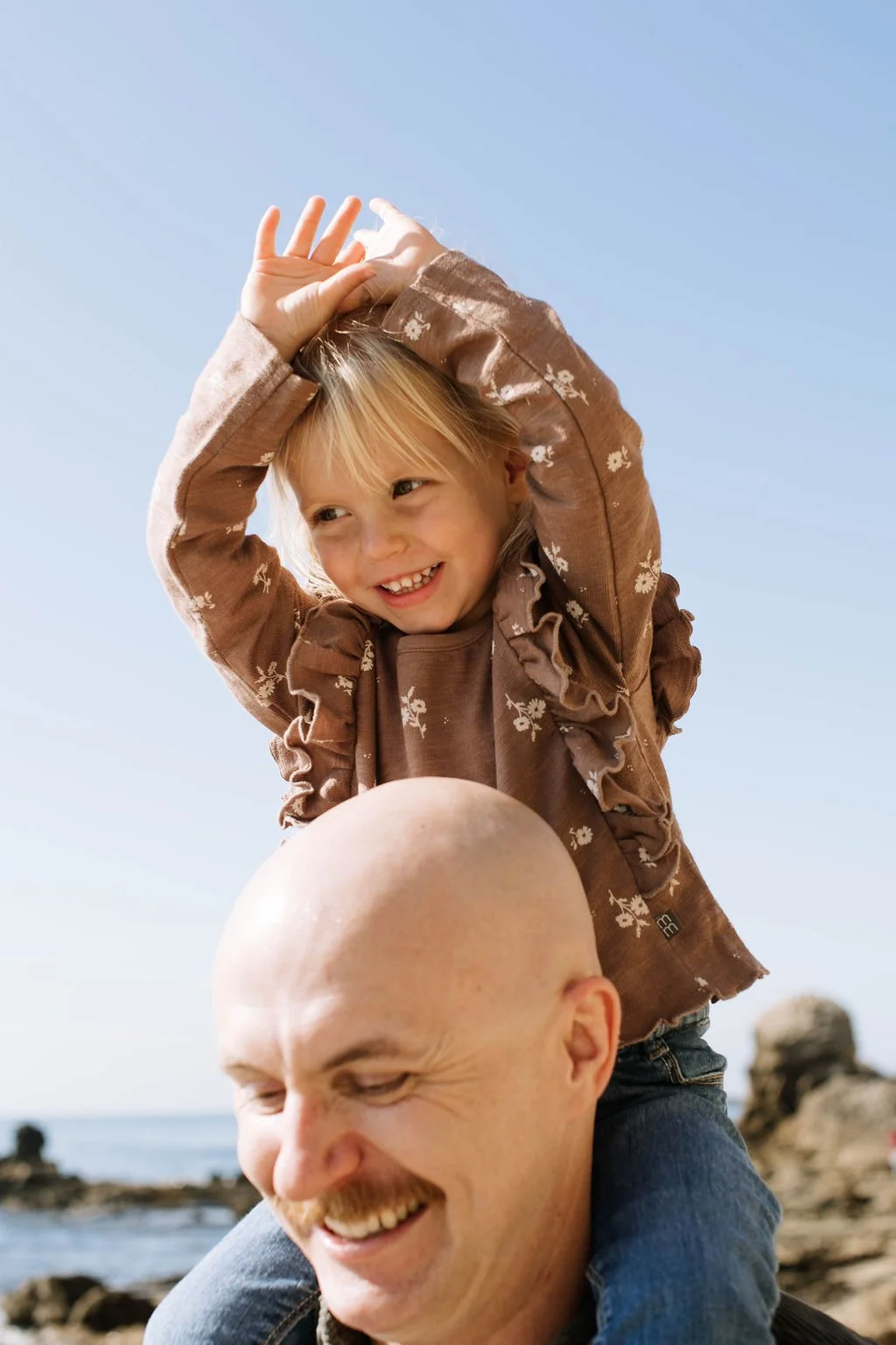 A young girl with blonde hair and a brown floral dress sitting on a man's shoulders, smiling and raising her hands above her head outdoors near the water with rocks in the background.
