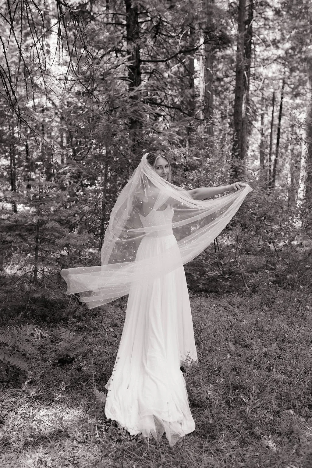 A woman in a white dress and veil poses in a forested area, holding her veil outstretched. The photo is in black and white.