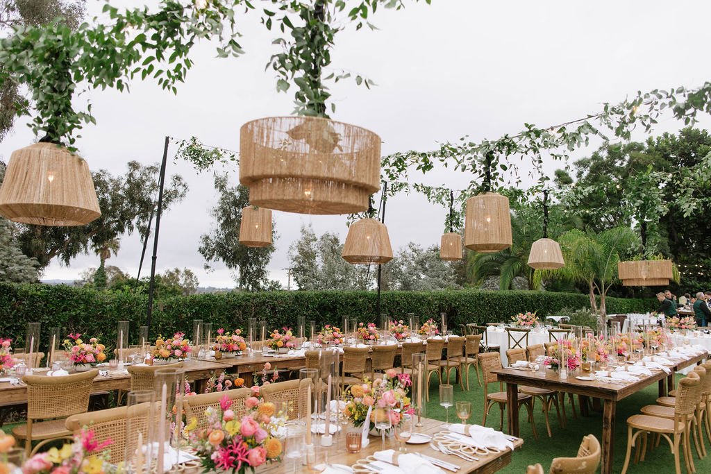 An outdoor event with long tables decorated with pink and peach floral arrangements, candles, and white table settings, under hanging wicker light fixtures and green leafy decorations.