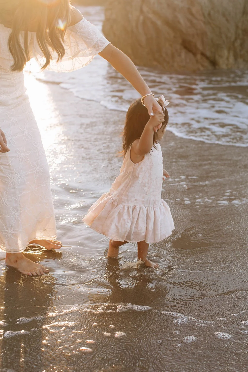 A woman holding a young girl’s hand while walking barefoot in shallow ocean water during sunset. The woman is wearing a white dress with embroidered floral patterns, and the girl is wearing a light pink dress. The scene is warm and peaceful with rock